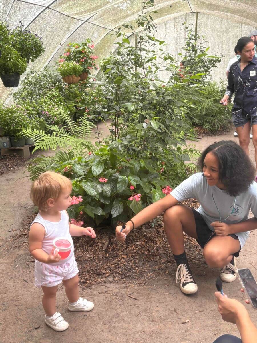 Abby kneeling beside a child among flowering plants inside the Chattahoochee Nature Center butterfly habitat
